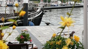 Un port de plaisance avec des bateaux amarrés, une enseigne sur laquelle on peut lire « ERNSTINE », et une table avec des fleurs au premier plan.