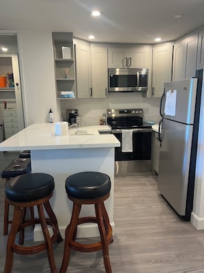 A modern kitchen with a central island, stainless steel appliances, and wooden stools. - Oceanview Retreat at North Coast Village B107 (Oceanside)