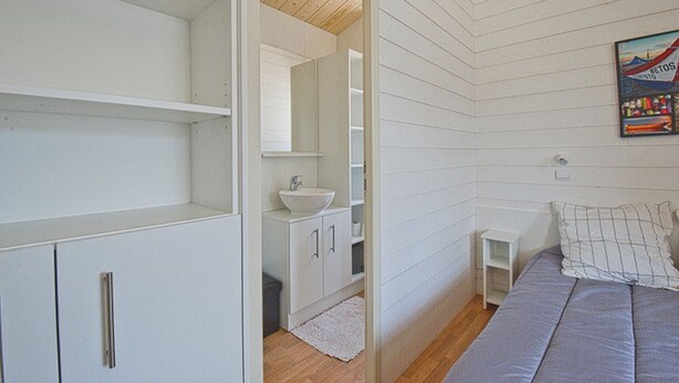 Quarto com cama, mesa de cabeceira, armário branco pequeno, espelho e teto de madeira.