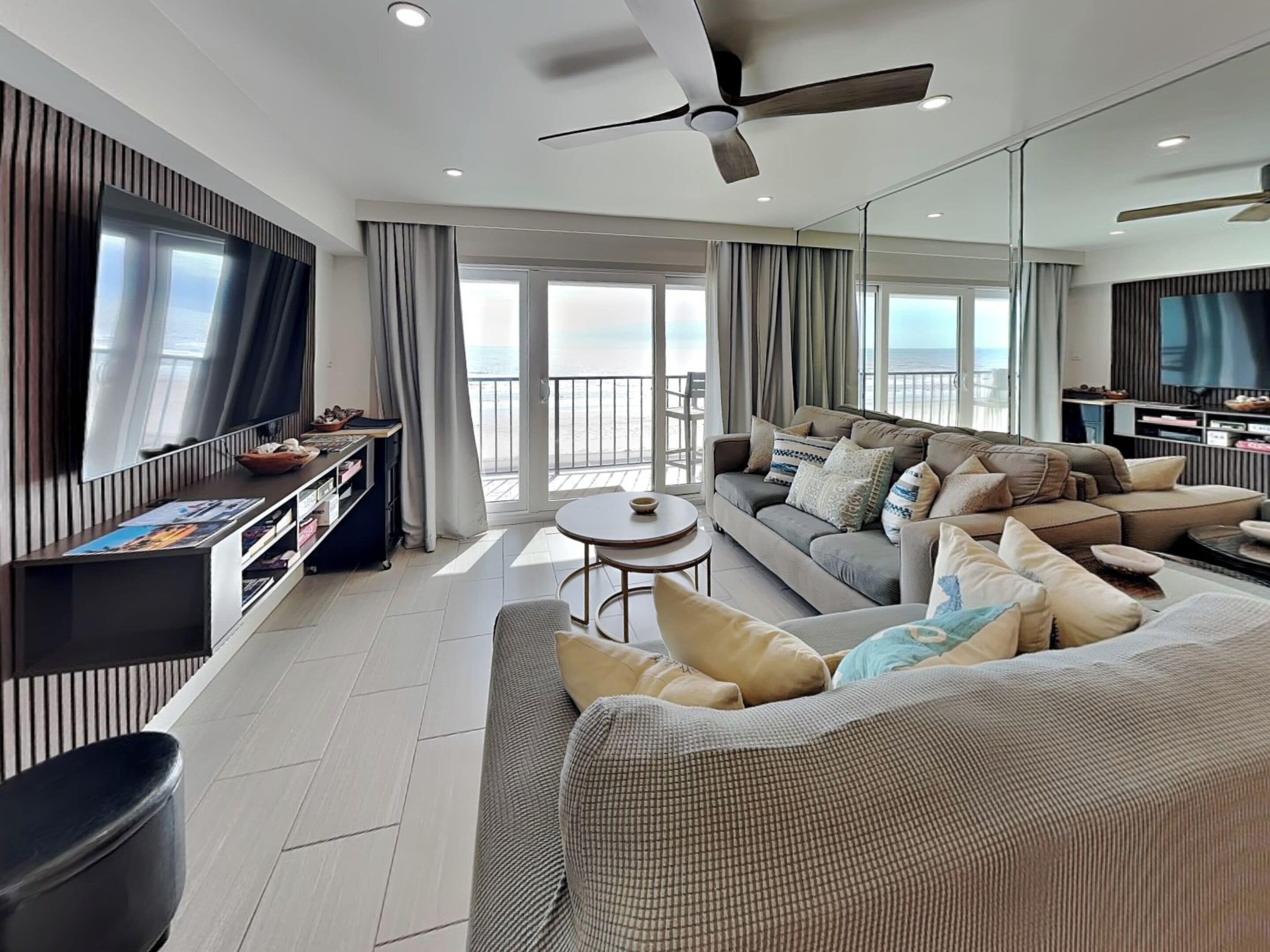 A modern living room with a sofa, a coffee table, and a TV. There is a balcony with a view of the ocean.