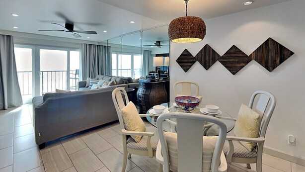 A modern living room with a sofa, dining table, and chairs. There is a ceiling fan and a decorative wall piece above the table. - By the Sea Resort 507-oceanfront View (Galveston)