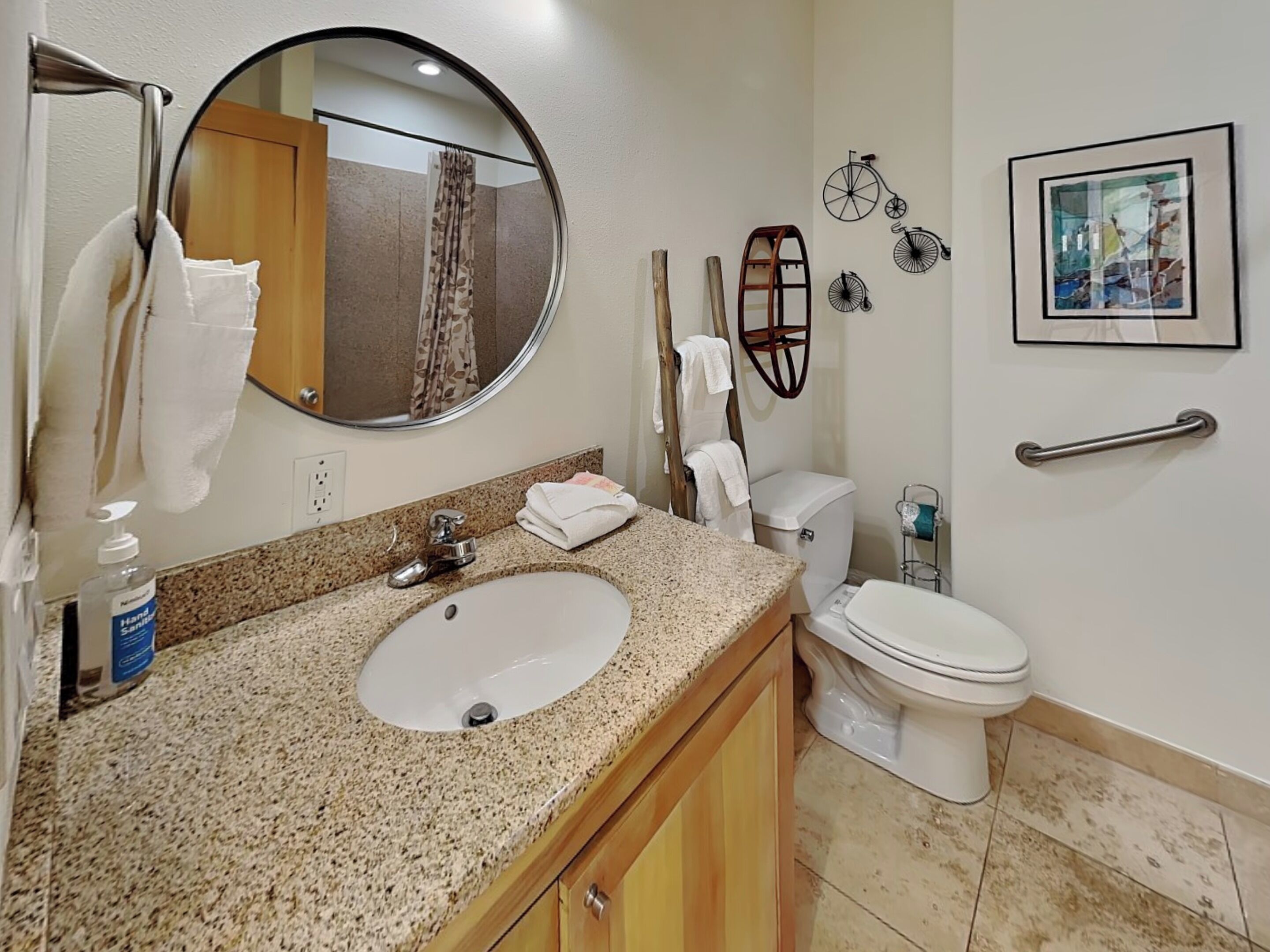 A bathroom with a granite countertop sink, a round mirror, a toilet, and a hand sanitizer dispenser.