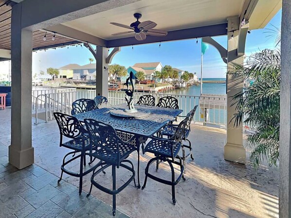 Une terrasse extérieure avec une table et des chaises, une sculpture décorative et une vue sur le bord de l’eau, avec des bâtiments et des palmiers.