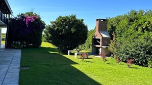 Une pelouse bien entretenue, agrémentée d’un espace barbecue et d’une cheminée en briques, le tout entouré d’une végétation luxuriante et sous un ciel d’un bleu éclatant.