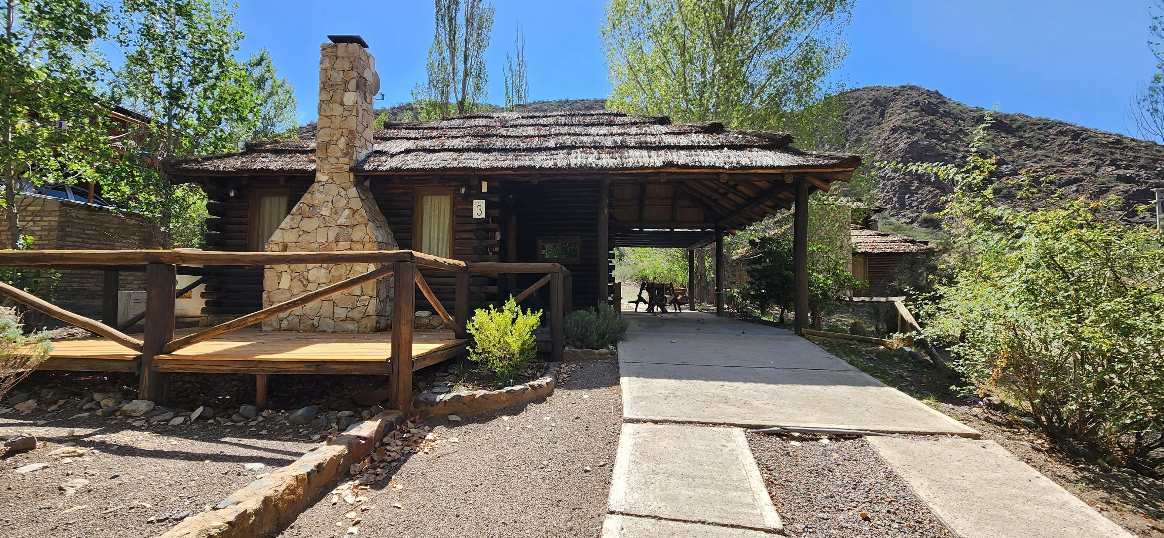 A rustic lodge with a stone chimney, wooden deck, and a pathway leading to a covered area.