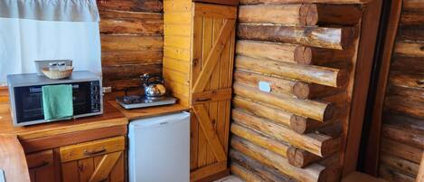 A compact kitchen with wooden walls, a microwave, a kettle, and a small refrigerator.