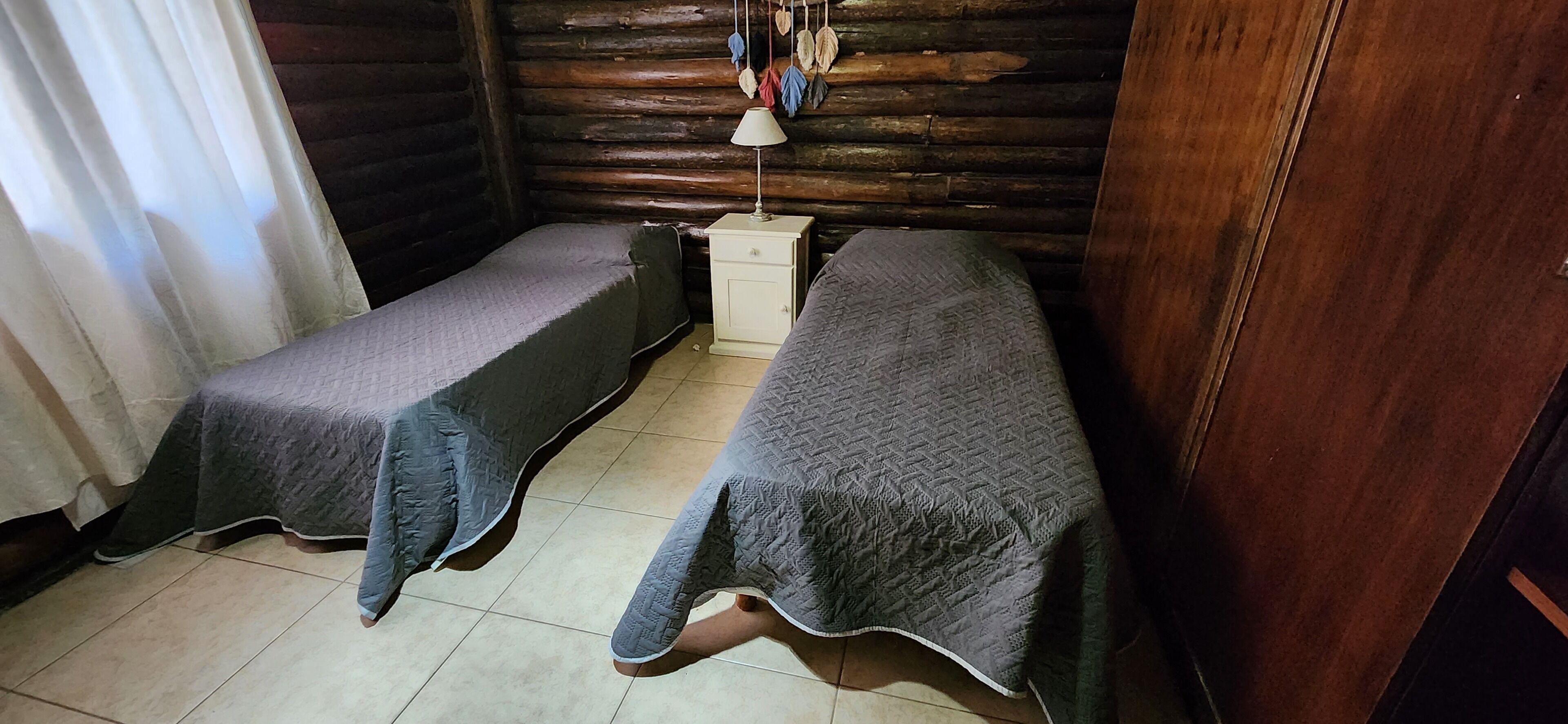 A small, cozy room with two single beds, a nightstand with a lamp, and a window with curtains.