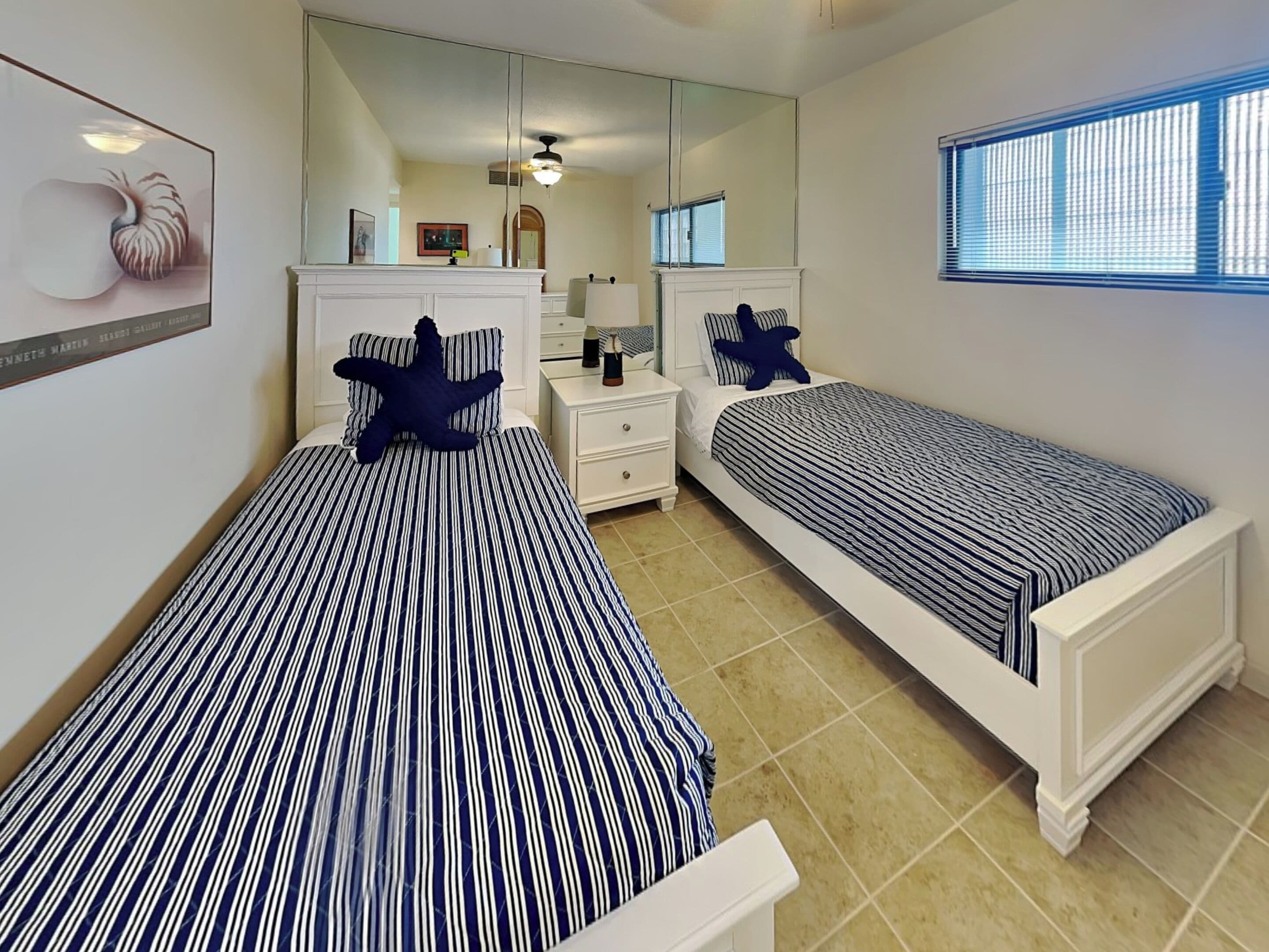 A bedroom with two beds, a large mirror, a window with blinds, and a tiled floor.