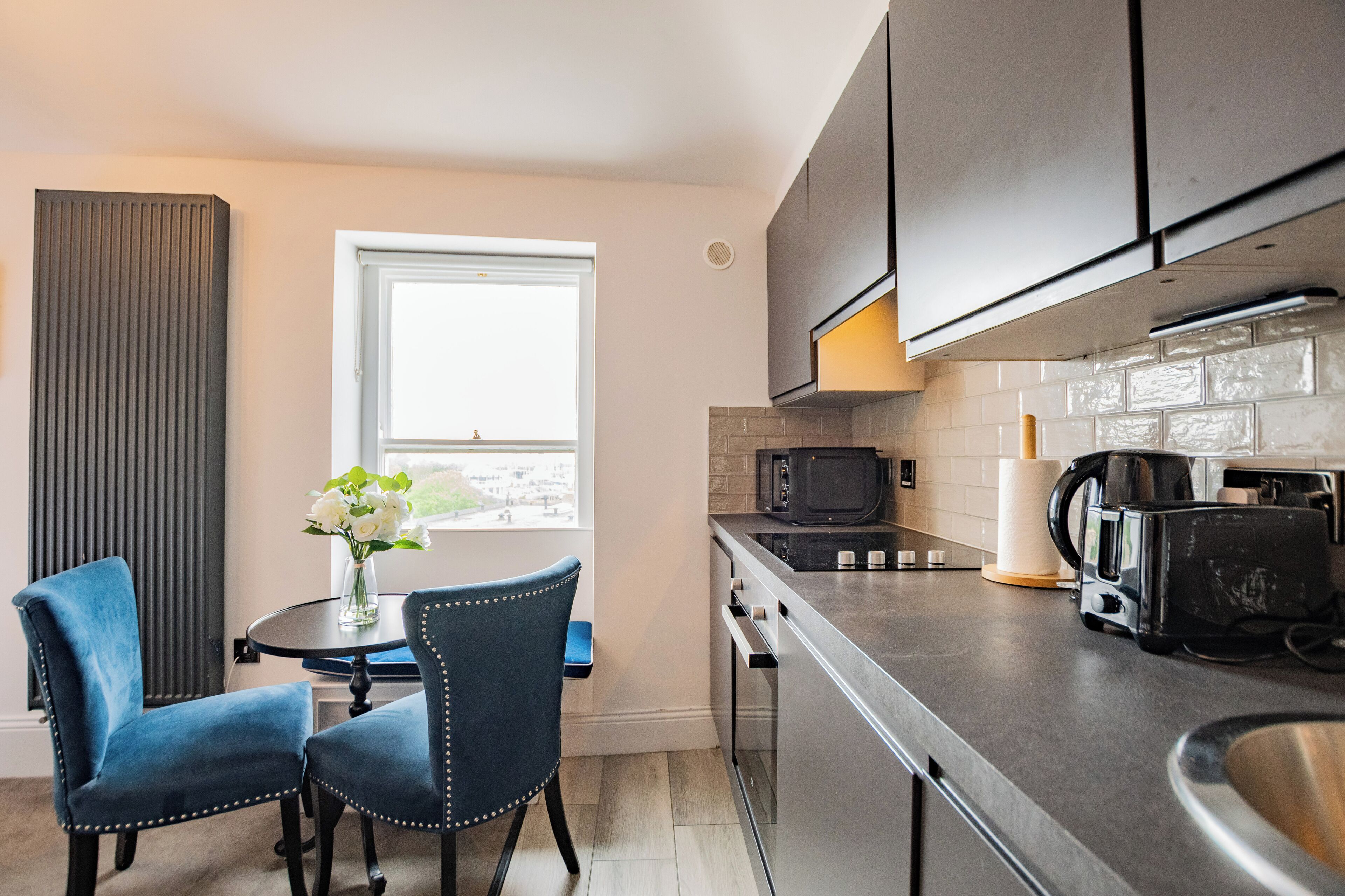 A modern kitchen with dark cabinets, a microwave, a coffee maker, and a toaster. There is a dining table with two chairs and a vase with flowers.