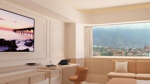 A modern hotel room with a large TV displaying a scenic view, a desk with a laptop, a comfortable sofa, and a view of a cityscape and mountains through the window. - Hotel Londres (Caracas)
