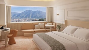 A modern hotel room with a large window offering a city and mountain view, a sofa, a bed, a small table, and a desk with a laptop. - Hotel Londres (Caracas)