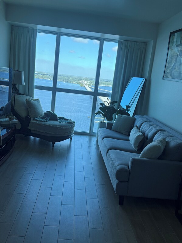 A room with a large window, a sofa, a chair, a lamp, and a mirror. - Luxury II | Breathtaking 24th-floor view of the Caloosahatchee River (fort myers)
