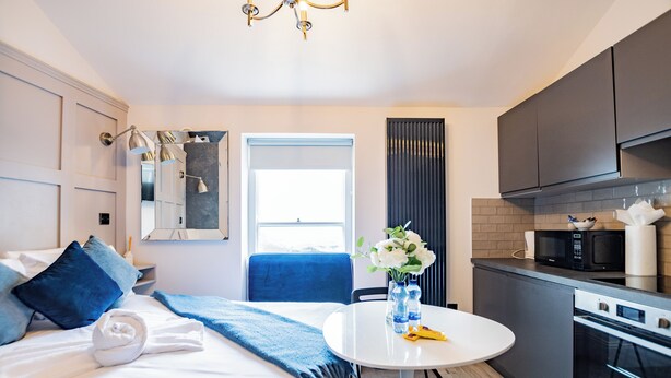 A modern hotel room with a bed, a round table with a vase of flowers, a microwave, and a built-in oven. - Superb Studio with Marina Views (Howth)