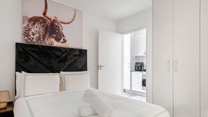 Ein modernes Schlafzimmer mit einem großen Bett, einem Kopfteil mit einem Kunstwerk zum Thema Stierkopf, einem weißen Kleiderschrank und Blick in den Küchenbereich.