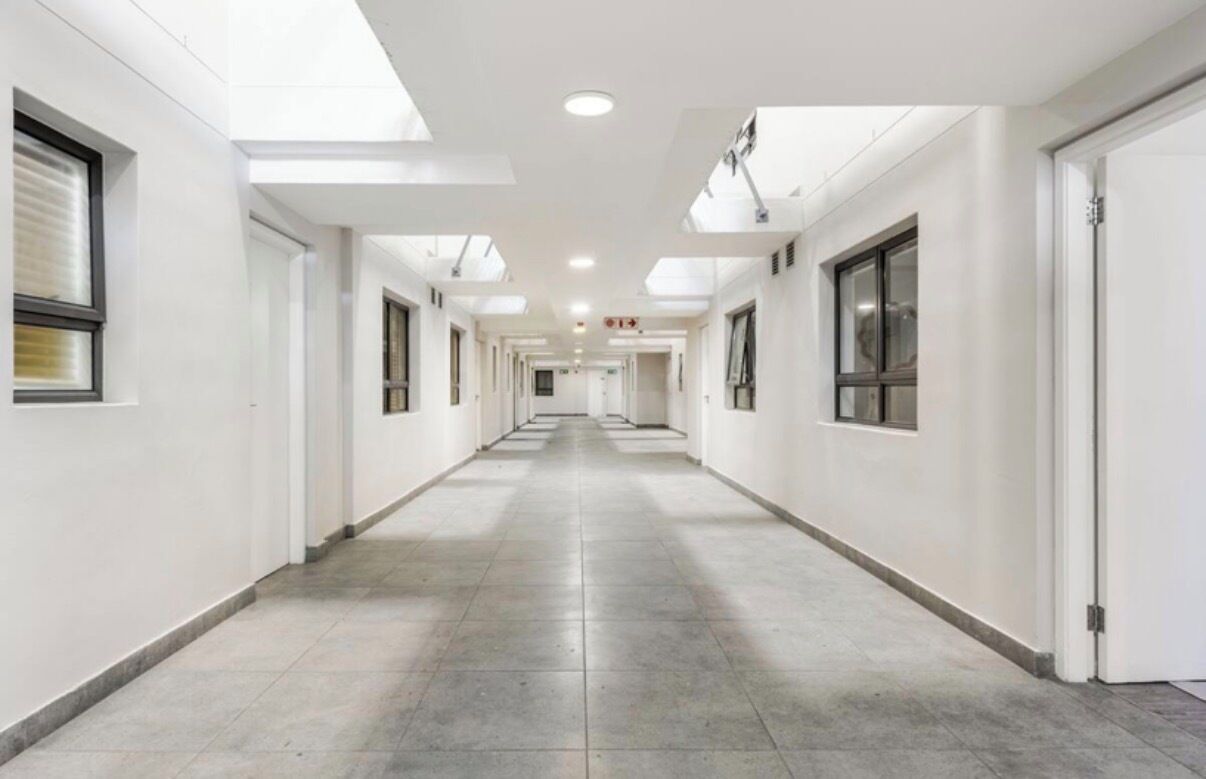 Um corredor comprido e estreito, com paredes brancas e portas em ambos os lados, que leva a uma área central com uma luminária no teto.