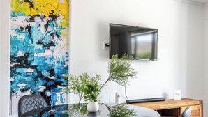 Ein moderner Essbereich mit Glastisch, schwarzen Stühlen, einer Holzkommode, einem großen abstrakten Gemälde und einem wandmontierten Fernseher.