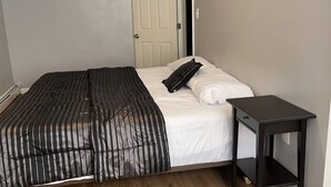 Un letto singolo con una coperta a righe nere e cuscini bianchi, un comodino nero piccolo e una porta chiusa sullo sfondo.