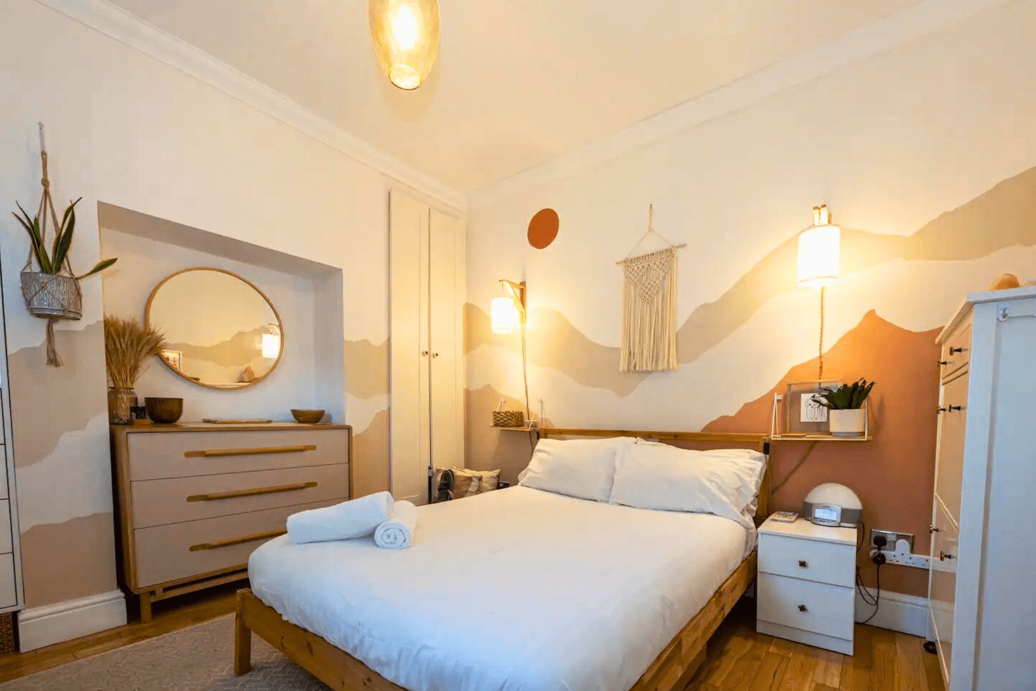 Une chambre à coucher comprenant un lit, des tables de chevet, une commode, un miroir, des lampes fixées au mur et un élément décoratif mural.