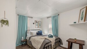 Ein Schlafzimmer mit einem Bett, Vorhängen, einem Nachttisch, einer kleinen Pflanze und einem Wandregal.