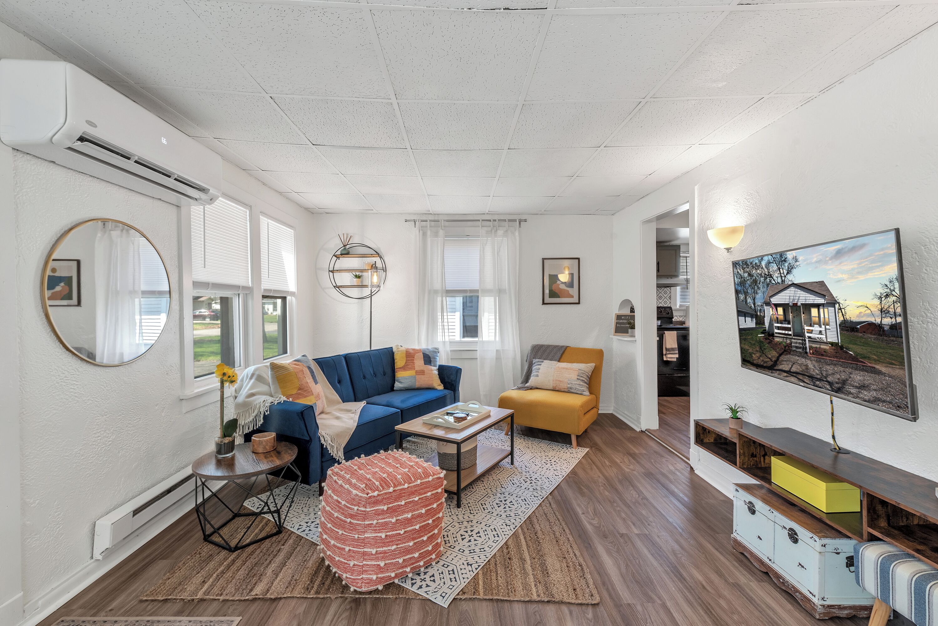 Un salon moderne avec un canapé bleu, un fauteuil jaune et une table basse. On y trouve un miroir rond, une petite table d’appoint et un tapis à motifs.