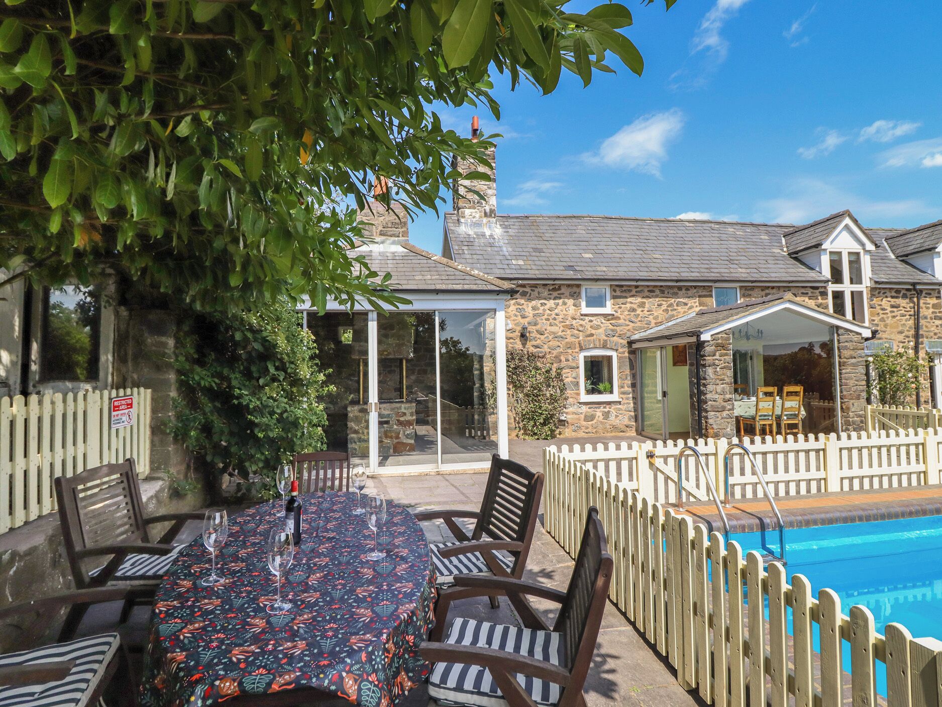 A poolside area with a table set for dining, a stone building with a glass enclosure, and a wooden fence surrounding the pool.