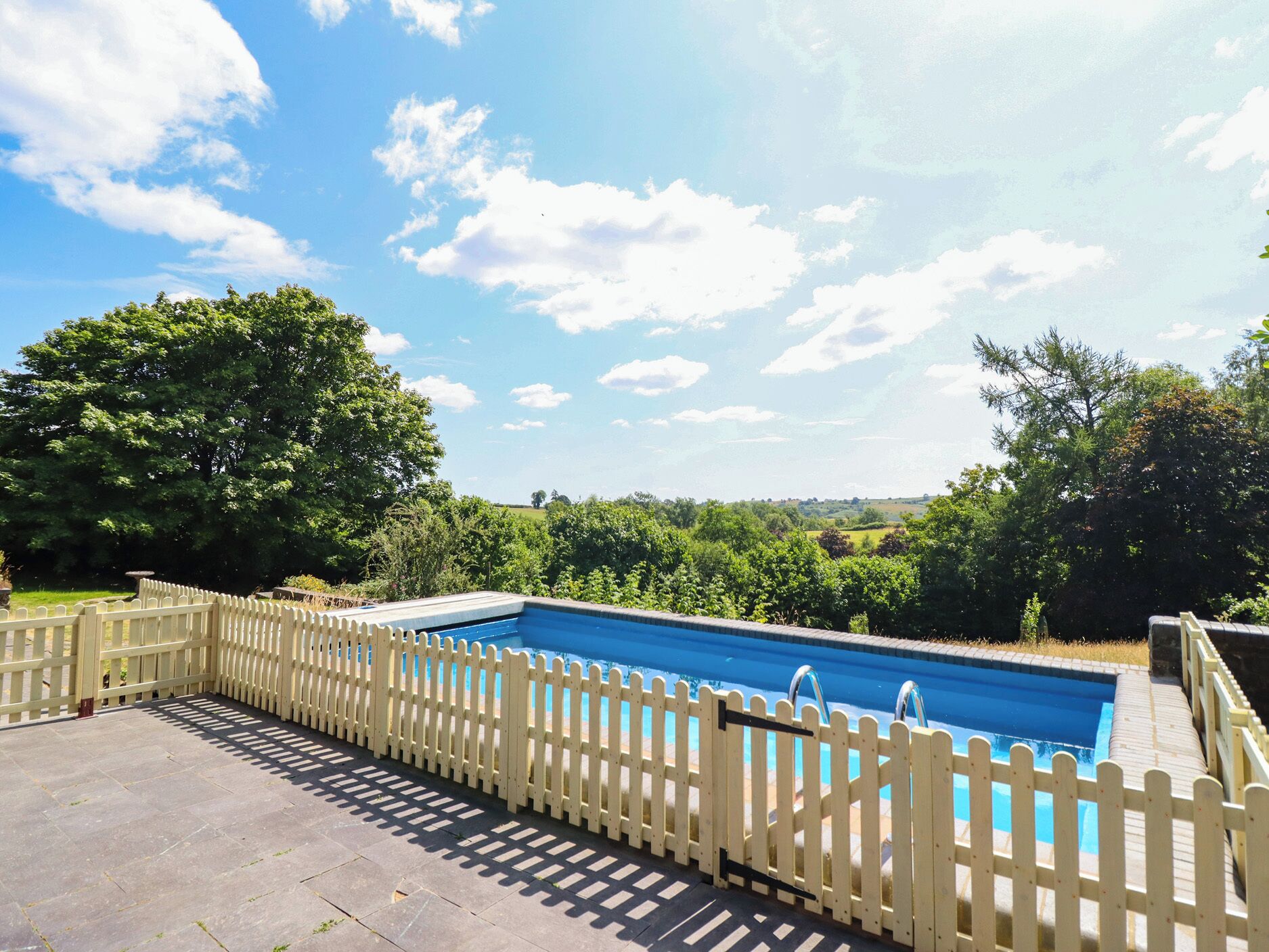 A swimming pool with a wooden fence, surrounded by a paved area and trees.