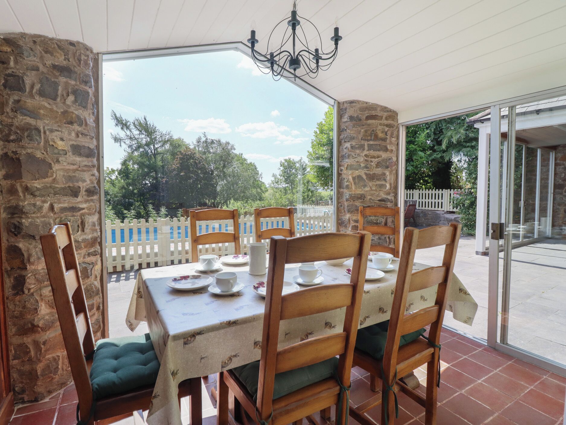A covered patio with a table set for four, stone walls, and a view of a white picket fence and trees.