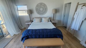 A bedroom with a large bed, two bedside tables with lamps, a wooden bench, a decorative wall hanging, and a window with curtains. - Absolute beach front comfort and luxury. (Waihi Beach)