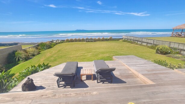 A wooden deck with two lounge chairs and a table, overlooking a grassy area and a beach. - Absolute beach front comfort and luxury. (Waihi Beach)