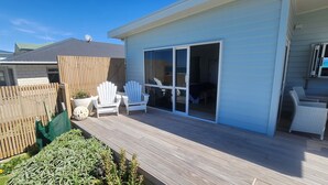 A covered deck with white Adirondack chairs, a sliding glass door leading to a patio, and a wooden fence. - Absolute beach front comfort and luxury. (Waihi Beach)