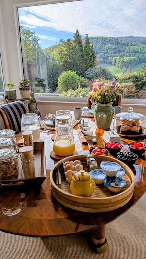 A breakfast table with various food items, a pitcher of orange juice, and a view of a forested landscape through a window.