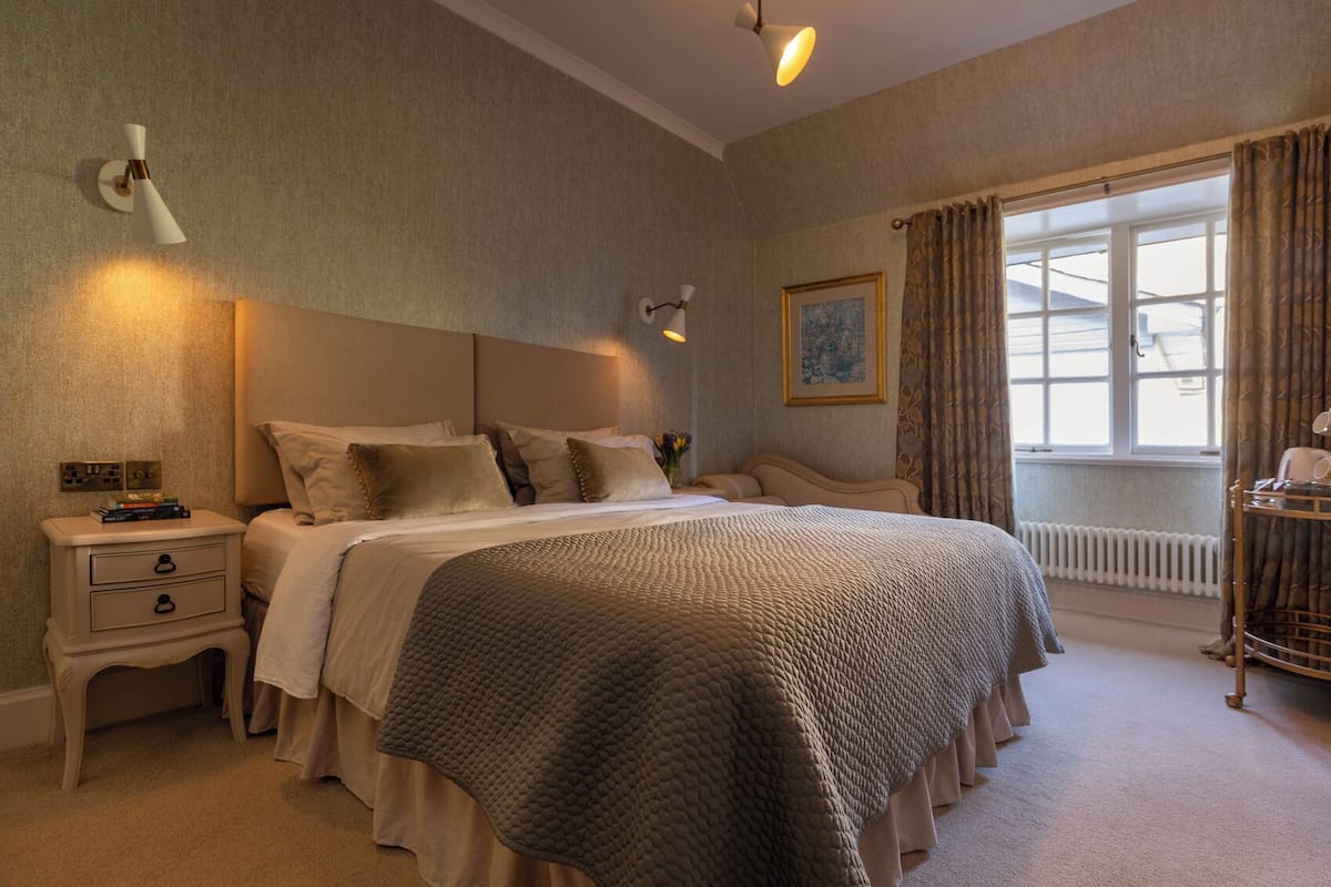 A neatly arranged bedroom with a large bed, bedside tables, a nightstand, a window with curtains, and a radiator.