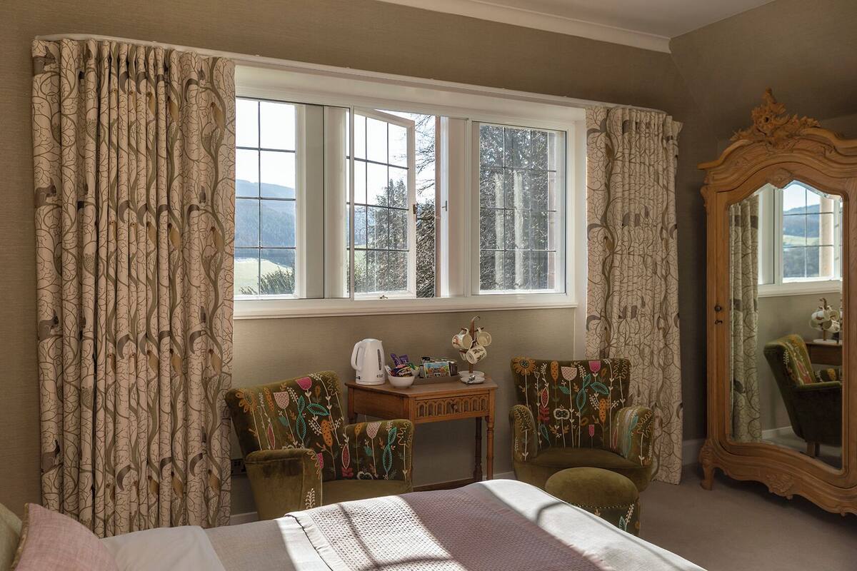 A room with a large window, a wooden dresser, two chairs, a small table with a kettle, and a mirror.