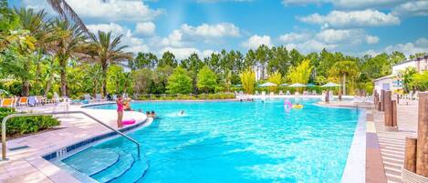 A swimming pool with clear blue water, surrounded by lounge chairs and umbrellas, with palm trees and a clear sky in the background.