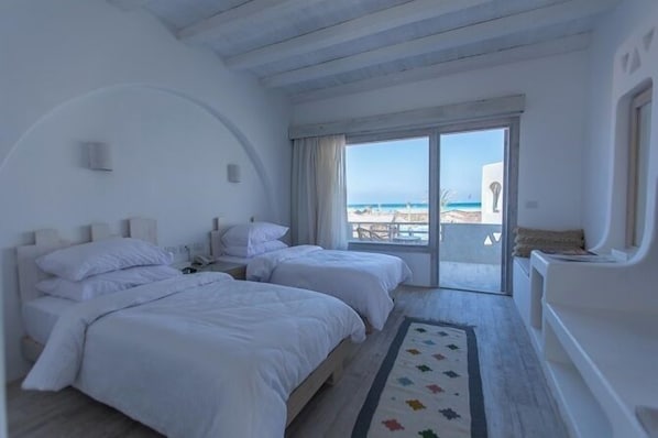 A room with two beds, a large window with a view of the beach, and a balcony with a table and chairs.