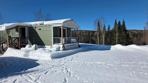 En liten stuga med en veranda, omgiven av snötäckt mark och träd.