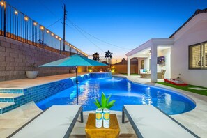 Una piscina con borde de azulejos, una mesa de madera con una lata y una botella, y un patio con una pared blanca y una ventana.