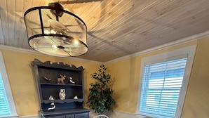 A dining area with a round wooden table, six chairs, a dark wooden cabinet with shelves, a potted plant, and a ceiling fan. - The Historical William Ellis Saint House (Bonavista)