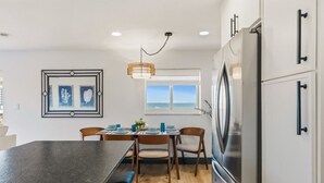 A modern kitchen with a central island, wooden chairs, a stainless steel refrigerator, and a window with a view of the sea. - Batiki West 609: Steps to Beach & Times Square, Gulf Views + Heated Pool! (Fort Myers Beach)