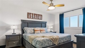 A bedroom with a bed, nightstands, a ceiling fan, a window with curtains, and a wall art piece. - Batiki West 609: Steps to Beach & Times Square, Gulf Views + Heated Pool! (Fort Myers Beach)