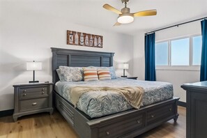 A bedroom with a bed, nightstands, a ceiling fan, a window with curtains, and a wall art piece. - Batiki West: Steps to Beach, Gulf Views + Pool! (Fort Myers Beach)