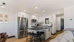 A modern kitchen with white cabinets, stainless steel appliances, and a central island with bar stools. - Batiki West 609: Steps to Beach & Times Square, Gulf Views + Heated Pool! (Fort Myers Beach)