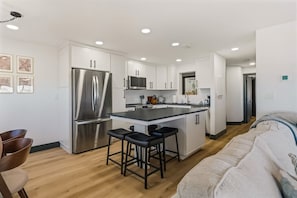 A modern kitchen with white cabinets, stainless steel appliances, and a central island with bar stools. - Batiki West: Steps to Beach, Gulf Views + Pool! (Fort Myers Beach)