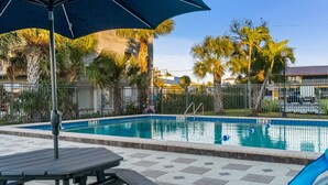A swimming pool with a tiled deck, surrounded by palm trees and a fence. - Batiki West 609: Steps to Beach & Times Square, Gulf Views + Heated Pool! (Fort Myers Beach)