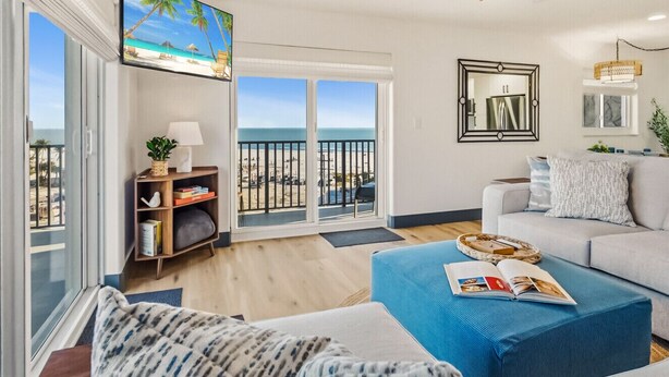 A modern living room with a large window overlooking a beach, a ceiling fan, and a sofa with a magazine on it. - Batiki West 609: Steps to Beach & Times Square, Gulf Views + Heated Pool! (Fort Myers Beach)