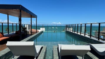 Uma piscina infinita com espreguiçadeiras, um espaço de estar coberto e vista para o oceano.
