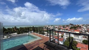 A rooftop pool with a wooden deck and glass railings, overlooking a coastal cityscape with buildings and palm trees. - Flat Comfort La Belle Formosa - Cabedelo (Cabedelo)