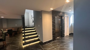 A modern hotel lobby with a staircase, a reception desk, and a sign indicating the elevator direction. - Flat Premium Legacy Suites - Cabo Branco (João Pessoa)