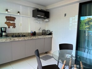 A modern kitchen with white tiled backsplash, a coffee maker, a gas stove, and a dining area with a glass table and chairs. - Flat Premium Sea View - Tabatinga Beach (Conde)