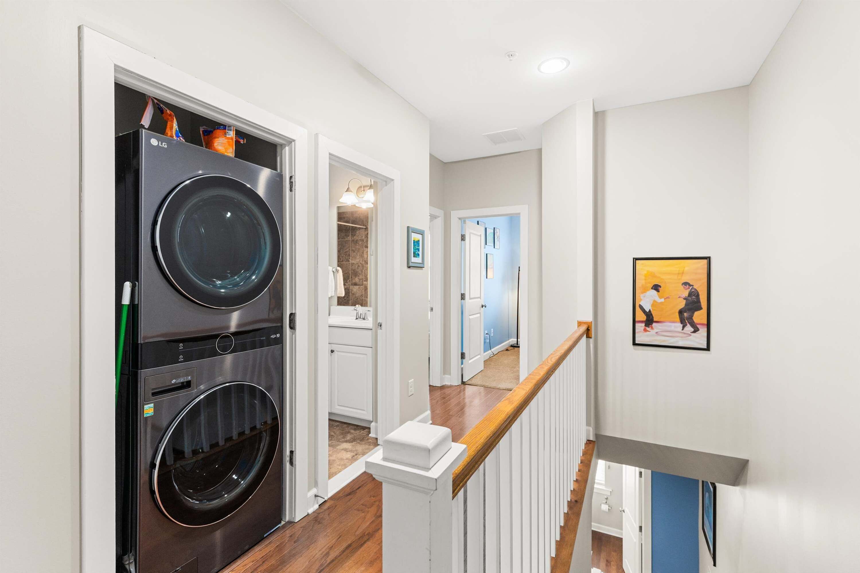 A laundry room with stacked washers and dryers, a wooden staircase, and a hallway leading to a bathroom.